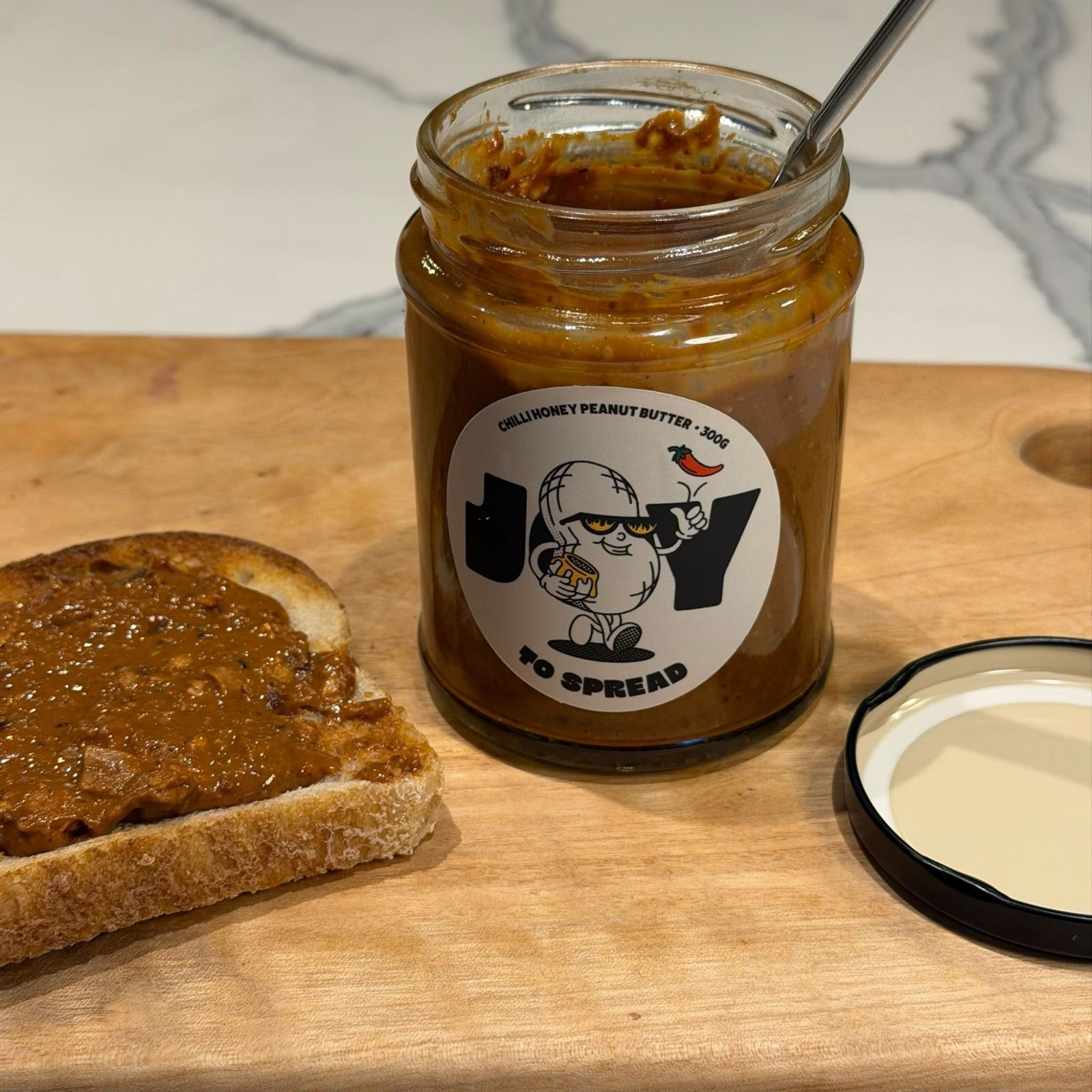 An open jar with a spoon resting in the thick spread, a slice of spread toast beside it